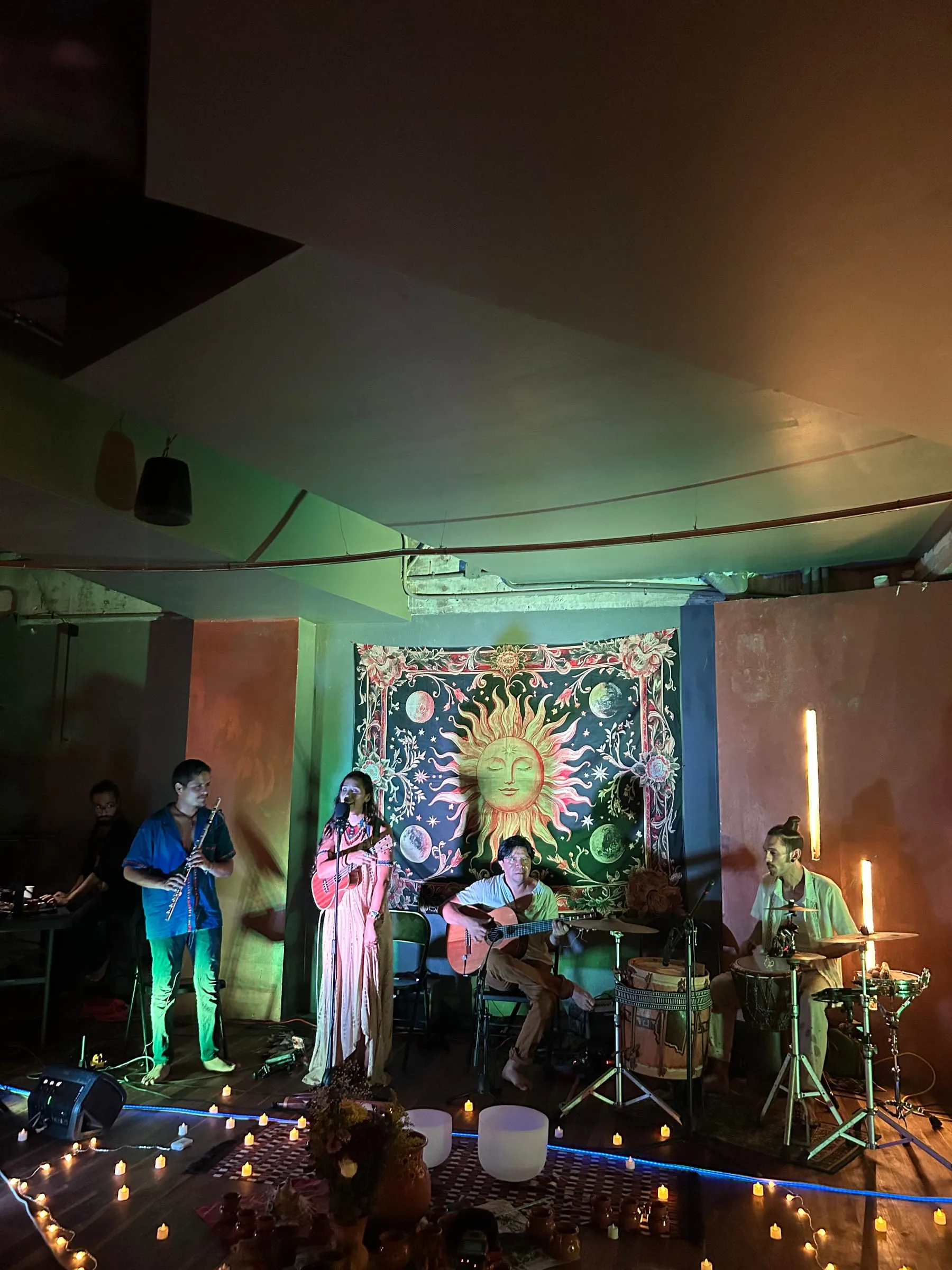 A live music session at Centro Calea: flute, voice, guitar and percussion in front of a sun tapestry, lit by candles.